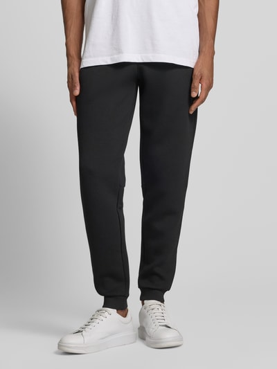 Männermodel trägt schwarze Sweatpants mit Viskose-Anteil Modell 'TRAIN PREMIUM SHIELD' von EA7 Emporio Armani, Elastischer-Material-Mix, Gerade Passform, Elastischer Bund, Reißverschlusstaschen, Gerippte-Abschlüsse, Ganzkörperansicht von vorne.