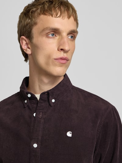 Męski model nosi Regular Fit Freizeithemd aus Cord mit Button-Down-Kragen von Carhartt Work In Progress, model 'Madison', czarna bawełna, szwy z logo, krój regular fit, kołnierz z guzikami, widok z przodu, zbliżenie na górną część ciała.