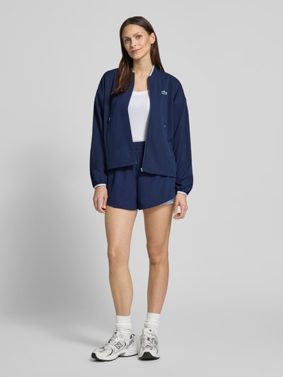 Frauenmodel trägt Regular Fit Trainingsjacke mit Baseball-Kragen von Lacoste Sport in Dunkelblau, unifarben mit Label Patch, Ganzkörperansicht von vorne.