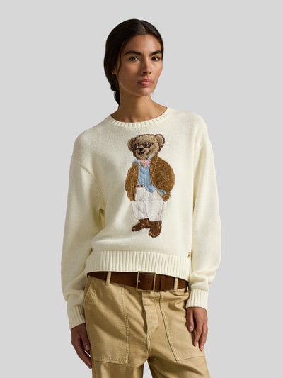 Modelka kobieca ma na sobie Regular Fit Strickpullover mit Polo Bear od Polo Ralph Lauren, kremowy, dzianinowa bawełna, krój regular fit, aplikacja Polo Bear, crew neck, długie rękawy zakończone ściągaczem, dół ściągaczowy, widok na górną część ciała z przodu.