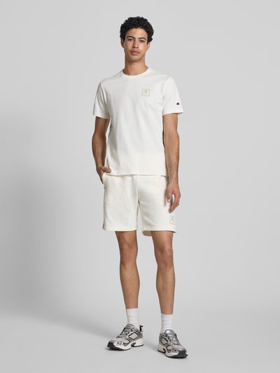 Männermodel trägt Shorts mit Logo und elastischem Bund von CHAMPION aus Baumwoll-Mix in Offwhite, Regular Fit, Ganzkörperansicht von vorne.