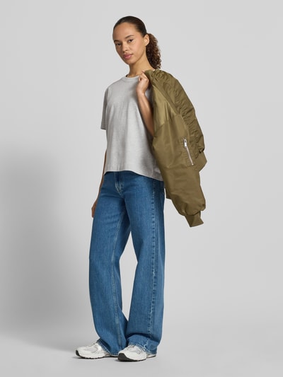 Modelka kobieca nosi szary damski t-shirt Boxy Fit T-Shirt aus Baumwoll-Viskose-Mix Modell 'ESSENTIAL' von Selected Femme, prosta faktura, szerokie niebieskie jeansy, białe sneakersy, detail kurtki w kolorze oliwkowym, damski t-shirt von Selected Femme, pełen widok z przodu.