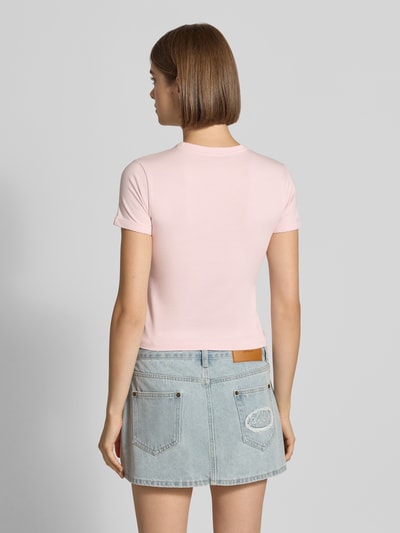 Frauenmodel trägt Slim Fit T-Shirt mit Strasssteinbesatz Modell 'Heart Baby' von GUESS Jeans, hellrosa, Rundhalsausschnitt, Ganzkörperansicht Rückansicht.
