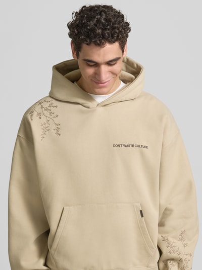 Männermodel trägt Oversized Hoodie aus Baumwoll-Mix von Don’t Waste Culture, Florale-Stickereien, Känguru-Tasche, elastische Abschlüsse, Rückenlänge 70 cm bei Größe M, Beige, Ganzkörperansicht von vorne.