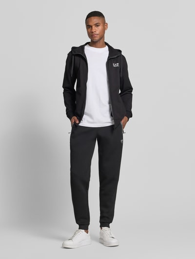 Männermodel trägt schwarze Sweatpants mit Viskose-Anteil Modell 'TRAIN PREMIUM SHIELD' von EA7 Emporio Armani, Gerade Passform, Reißverschlusstaschen, Gerippte Abschlüsse, Elastischer Bund, Elastischer-Material-Mix, Ganzkörperansicht von vorne.