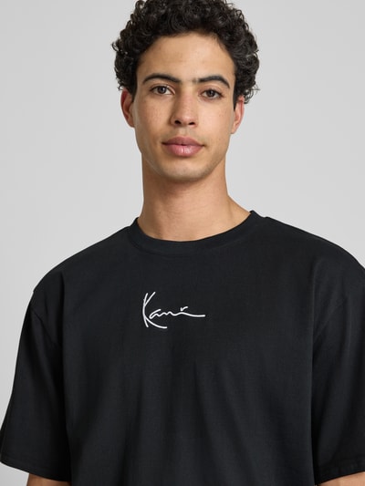 Męski model nosi T-Shirt mit Label-Stitching und Rundhalsausschnitt von KARL KANI, czarna bawełna, oversize, haft, zbliżenie na górną część ciała.
