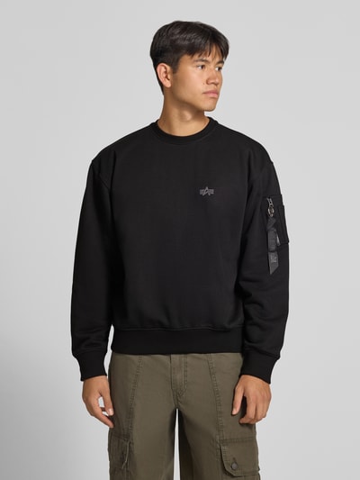 Męski model nosi bluza 'Sweatshirt mit Label-Details' od Alpha Industries, mieszanka bawełny, luźny krój, okrągły dekolt, detal z logo, kieszeń na rękawie z aplikacją z metką, prążkowane wykończenia, długość tyłu w rozmiarze M: 66 cm, długość rękawa w rozmiarze M: 62 cm, kolor czarny, widoczna gładka tekstura materiału, pełen widok z przodu.