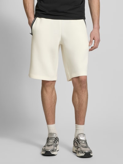 Męski model nosi Shorts mit elastischem Bund od EA7 Emporio Armani, kolor kremowy, materiał z domieszką wiskozy, krój regular fit, Szerokość w rozmiarze M: 41 cm, długość nogawki od wewnątrz: 28 cm, widok całej sylwetki z przodu.