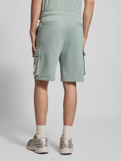 Männermodel trägt Sweatshorts mit Cargotaschen und Logo von CHAMPION in Mintgrün, Baumwoll-Mix, Relaxed Fit, Ganzkörperansicht Rückansicht.