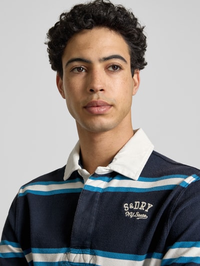 Männermodel trägt Regular Fit Poloshirt mit Logo-Stitching von Superdry, reine Baumwolle, dunkelblau mit weißen und blauen Streifen, Nahaufnahme Oberkörper.