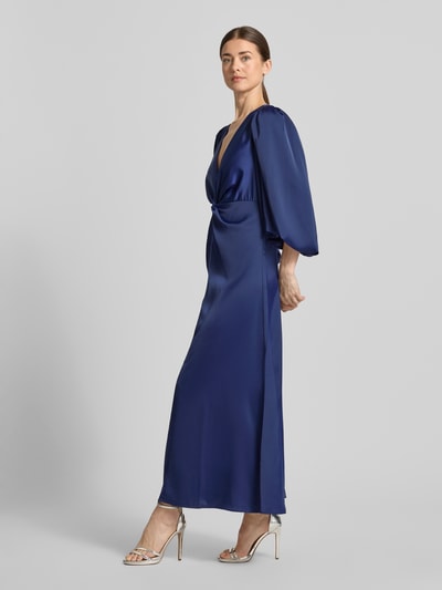 Vrouwenmodel draagt donkerblauwe maxi-jurk van YAS, materiaalmix met stretch, getailleerd model, V-hals, knoopdetail, ballonmouwen, model 'THEA', zijaanzicht, bijna volledige lichaamsopname.