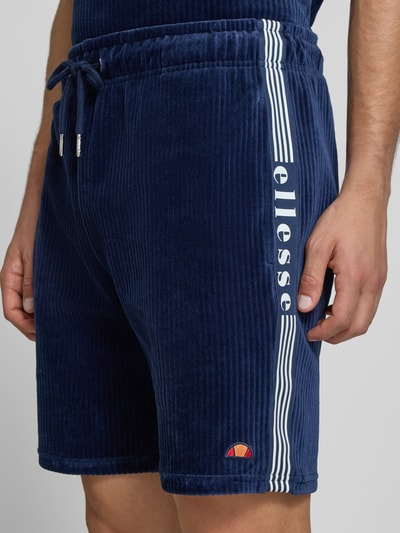 Mężczyzna nosi Shorts aus Cord Modell 'TOMATRO' von Ellesse, granatowy sztruks, elastyczny pas z tunelem, krój regular fit, bawełna ze stretchem, napis z boku, szerokość w rozmiarze M: 42 cm, długość nogawki wewnętrznej: 21 cm, zbliżenie na górną część nóg