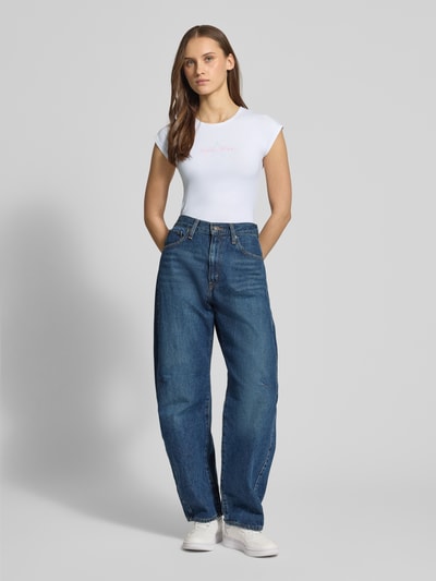 Frauenmodel trägt Slim Fit T-Shirt mit Logo-Stitching von Guess Jeans aus elastischem Baumwoll-Mix, Weiß, Rundhalsausschnitt, Ganzkörperansicht von vorne.