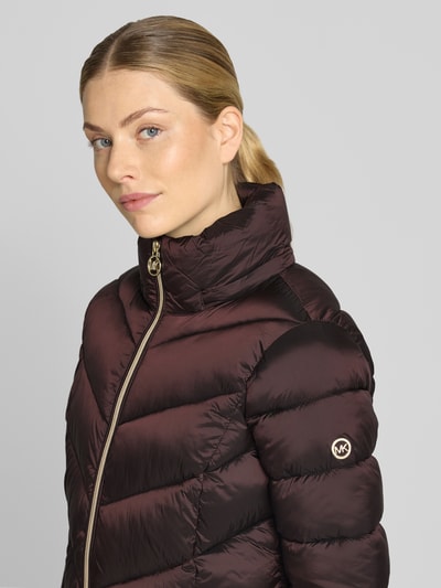 Frauenmodel trägt Steppmantel mit Kapuze und Reißverschlusstaschen von MICHAEL Michael Kors in dunklem Braun, Nahaufnahme Oberkörper. Gerader Schnitt, Stehkragen, seitliche Reißverschlusstaschen, glänzende Oberfläche, dezentes Markenlogo am Arm.