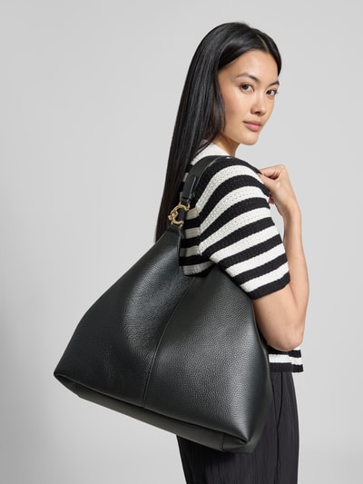 Vrouwenmodel draagt Tote Bag met Logo-Detail van Coccinelle in zwart met grof korrelige textuur, Hoofdvak met magneetsluiting, Schouderband, uitneembare binnenzak, Breedte 25 cm, Lengte 31 cm, Diepte 15 cm, bandlengte 54 cm, Ganzekörperaanzicht achterkant.
