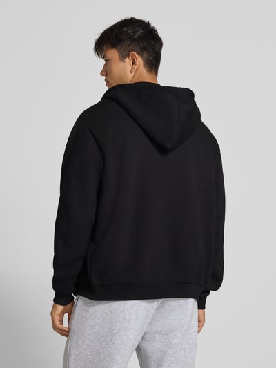 Männermodel trägt Sweatjacke mit Label-Stitching und Känguru-Tasche von Karl Kani im Baumwoll-Mix, Schwarz, Regular Fit, Ganzkörperansicht Rückansicht.