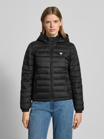Vrouwenmodel draagt Gewatteerd jack met capuchon van Guess Jeans, zwart, regular fit, ritssluiting, labeldetail, ritszakken. Ganzkörperansicht van voren. Ruglengte maat S: 60 cm, schouderbreedte: 45 cm, borstwijdte: 38 cm, mouwlengte: 62 cm.