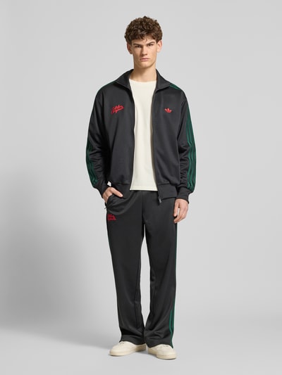 Männermodel trägt 'Sweatjacke mit Logo-Stitching und Stehkragen' van adidas Originals, zwart met groene accenten, materiaal-mix met katoen, recht model, opstaande kraag, ritssluiting, logo-stitching, zijritszakken, elastische boorden, voorkant volledige lichaamsopname.