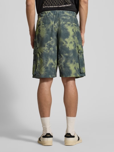Männermodel trägt Shorts mit Cargotaschen Modell 'Ethan' von Wood Wood aus Baumwoll-Mix, Relaxed-Fit, grün-blau gemustert, Ganzkörperansicht Rückansicht. Bundweite bei Größe M: 41 cm, Innenbeinlänge bei Größe M: 20 cm.