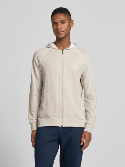 Männermodel trägt Regular Fit Sweatjacke aus Baumwoll-Mix von BOSS in Beige mit Kapuze, Kontraststreifen, Reißverschluss, Ganzkörperansicht von vorne.