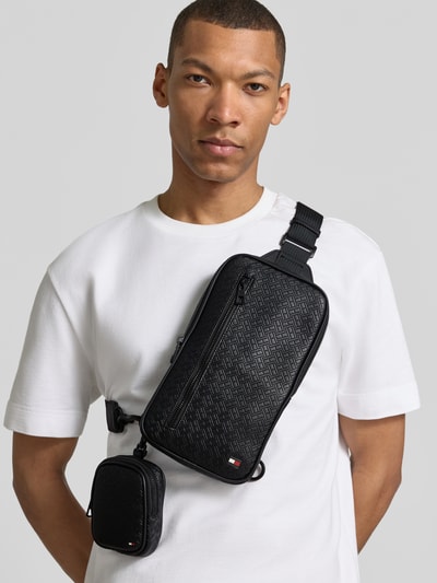 Männermodel trägt torba na ramię 'Umhängetasche mit Logo-Detail Modell 'EMBOSSED FLAG SLING' von Tommy Hilfiger z imitacją skóry, wzór flag-monogram-prägung, detal z logo, pasek na ramię, główna przegroda, przegródka zapinana na zamek błyskawiczny, przegródka z siateczki, krój prostokątny, kolor czarny, wykończenie z sztucznej skóry, ganzkörperansicht z przodu.