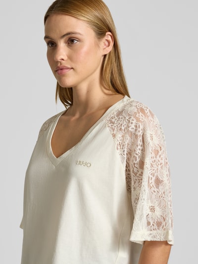 Frauenmodel trägt T-Shirt mit Spitze und Logo-Stitching von LIU JO SPORT in Creme mit V-Ausschnitt und Raglanärmel, Nahaufnahme Oberkörper.