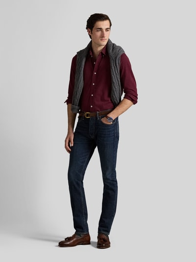 Mężczyznamodel nosi Regular Fit Freizeithemd aus reiner Baumwolle von Polo Ralph Lauren w kolorze bordowym, ciemnoniebieskie jeansy, brązowe buty, szary sweter na ramionach, widoczne faktury tkanin, widok całej sylwetki z przodu.