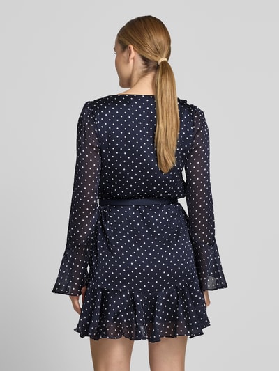 Vrouwenmodel draagt Minikleid met Rüschenbesatz van GUESS in donkerblauw met witte stippen, getailleerd, met ruches, met lange mouwen, V-hals, afneembare tailleriem, ruglengte maat S 83 cm, borstwijdte 42 cm, mouwlengte 62 cm. Ganzkörperansicht Rückansicht.