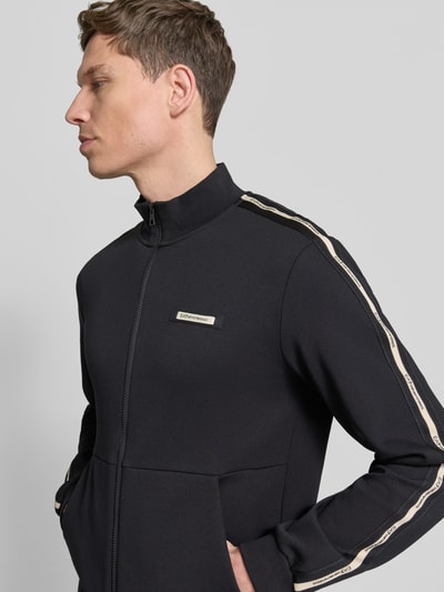 Męski model nosi bluzę rozpinaną z stójką i zamkiem błyskawicznym dwustronnym od EA7 Emporio Armani, krój regular fit, wpuszczane kieszenie, detal z logo, długość tyłu w rozmiarze M: 68 cm, długość rękawów w rozmiarze M: 67 cm, kolor czarny z beżowymi paskami, tekstura gładka, detal metalowy, widok na górną połowę ciała z przodu.