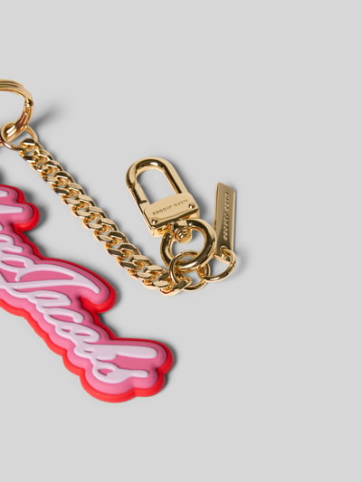 Produktfoto, Charm mit Logo-Detail von Marc Jacobs in Ketten-Optik aus goldfarbenem Metall mit Karabinerhaken, Cursive-Optik und Logo-Prägung auf hellgrauem Hintergrund.