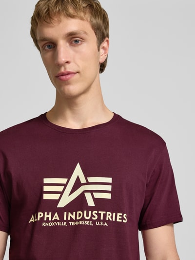 Męski model nosi T-Shirt mit Logo und Rundhalsausschnitt von Alpha Industries, bawełna, krój regular fit, logo, kolor bordowy, widok na górną część ciała z przodu.