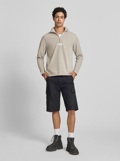 Mężczyzna model nosi Fleecepullover mit Reißverschluss Modell 'GLACIER' von The North Face, krój regular fit, stójka, wyhaftowane logo, zamek błyskawiczny, długość tyłu w rozmiarze M: 70 cm, długość rękawów w rozmiarze M: 67 cm, jasnobeżowy i czarny, gładka tekstura, widok całej sylwetki z przodu.