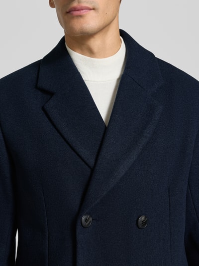 Männermodel trägt Regular Fit Mantel mit Reverskragen von Lindbergh in Dunkelblau, strukturierte Oberfläche, Nahaufnahme Oberkörper, KI-modifiziertes Bild. Rückenlänge bei Größe M: 94 cm, Schulterbreite: 45 cm, Brustweite: 52 cm, Ärmellänge: 66 cm, doppelte Knopfleiste, Pattentaschen.