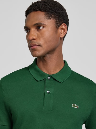 Männermodel trägt Slim Fit Poloshirt aus reiner Baumwolle von Lacoste in Dunkelgrün, glatte Textur, Nahaufnahme Oberkörper.