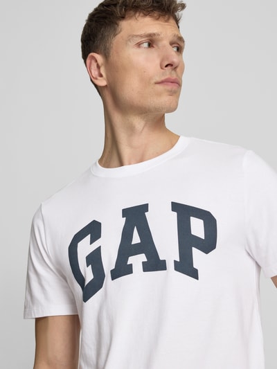 Männermodel trägt T-Shirt mit Logo und Rundhalsausschnitt von GAP aus reiner Baumwolle im Regular-Fit, weiß mit schwarzem Logo, Nahaufnahme Oberkörper.