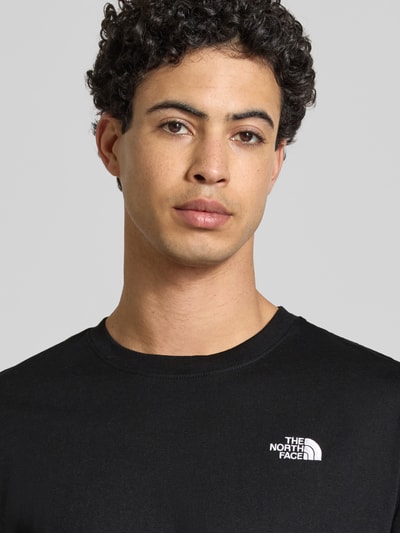 Mężczyzna nosi Regular Fit T-Shirt mit Logo-Stitching von The North Face, czarny kolor, okrągły dekolt, bawełna, krój regular fit, zbliżenie na górną część ciała.