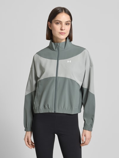 Frauenmodel trägt Trainingsjacke mit Stehkragen und Eingrifftaschen von Under Armour in Grün und Grau, leicht glänzende Textur, Regular-Fit, Logo, Reißverschluss, Schulterbreite 62 cm, Rückenlänge 60 cm, Brustweite 57 cm, Ärmellänge 50 cm, Ganzkörperansicht von vorne.