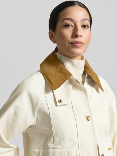 Kobiece model nosi Regular Fit Parka aus reiner Baumwolle od Barbour, efekt: jednolity kolor, krój regular fit, wykładany kołnierz, zapięcie na zamek błyskawiczny oraz listwę z zatrzaskami, aplikacja z wyhaftowanym logo, długi rękaw, kieszenie z patką, kieszeń wewnętrzna, materiał: bawełna, zbliżenie na górną część ciała