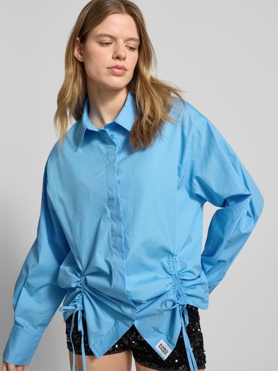 Frauenmodel trägt Oversized Bluse mit Kordelzug und verdeckter Knopfleiste von Karo Kauer aus Reine Baumwolle, Oversized-Fit, Hemdblusenkragen, Sportmanschetten, Bund mit doppeltem Kordelzug und Bindeverschluss, Label-Patch, Rückenlänge 75 cm, Ärmellänge 61 cm, einfarbiges Hellblau mit glatter Textur, Ganzkörperansicht von vorne.