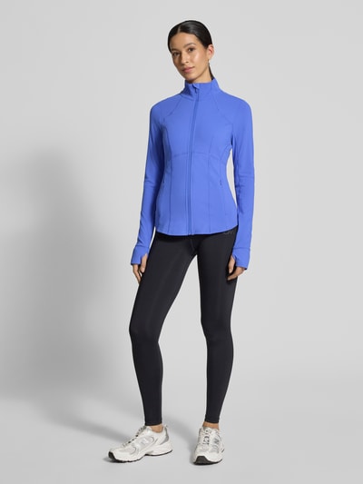 Frauenmodel trägt Slim Fit Jacke mit Stehkragen Modell 'Storm' von Fabletics in Blau mit Reißverschluss und Daumenschlaufen, Ganzkörperansicht von vorne.