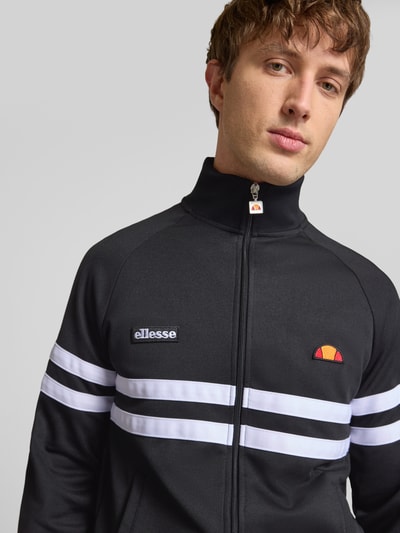Męski model nosi bluza rozpinana z mieszanki bawełny Ellesse, model 'RIMINI', czarna z białymi paskami, prosta krój, stójka, zamek błyskawiczny, detale z logo, elastyczne wykończenia, długość tyłu 71 cm, długość rękawów 43 cm, widok na górną część ciała z przodu.