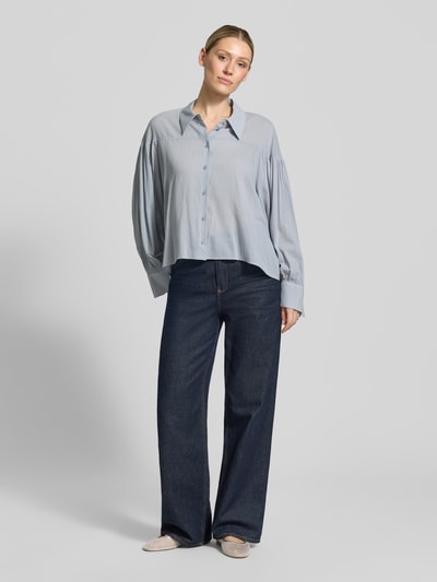 Frauenmodel trägt Relaxed Fit Bluse Modell 'SELENI' von Drykorn aus Baumwoll-Viskose-Mix, hellblau mit Hemdblusenkragen und Knopfleiste, Ganzkörperansicht von vorne. Rückenlänge bei Größe 36: 59 cm, Ärmellänge 63 cm.