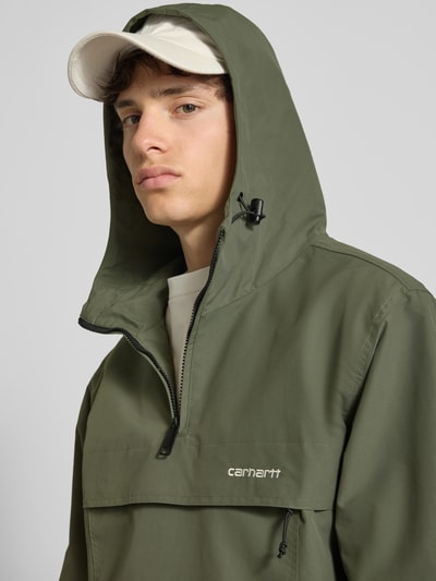 Männermodel trägt Windbreaker mit Label-Stitching von Carhartt Work In Progress in Olivgrün, Reißverschluss, Kapuze, Nahaufnahme Oberkörper, Herren Regular-Fit, Rückenlänge 73 cm, Schulterbreite 48 cm, Brustweite 55 cm, Ärmellänge 66 cm, Größe M.