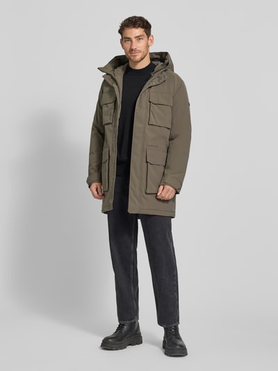 Męski model nosi Parka mit Pattentaschen Modell 'AIDEN' od Didriksons, oliwkowy kolor, regular fit, długość do kolan, widok całej sylwetki z przodu. Regularny krój, kaptur, kieszenie z patkami, pasek na rękawie, długość tyłu 92 cm, szerokość ramion 47 cm, szerokość klatki piersiowej 56 cm, długość rękawa 69 cm.