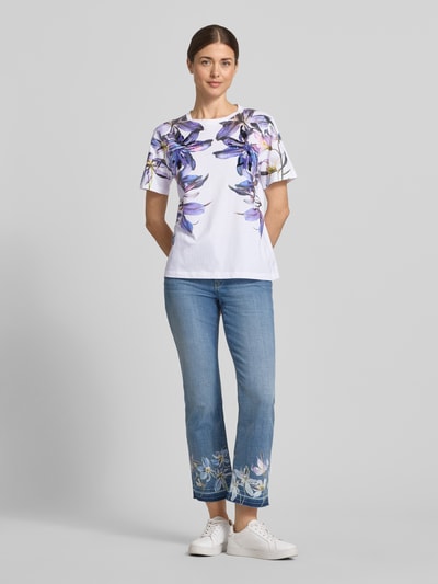 Frauenmodel trägt Damen T-Shirt im Allover-Look mit Pailletten von Marc Cain aus Reine Baumwolle, Regular-Fit, weiß mit violetten Blumen, Rundhalsausschnitt, Rückenlänge 59 cm bei Größe 36, Ganzkörperansicht von vorne.