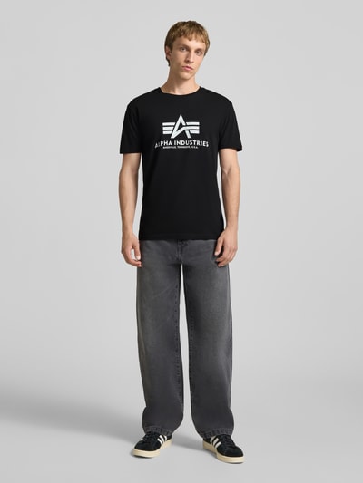 Männermodel trägt T-Shirt mit Logo und Rundhalsausschnitt von Alpha Industries, schwarze Baumwolle, Regular Fit, Ganzkörperansicht von vorne.