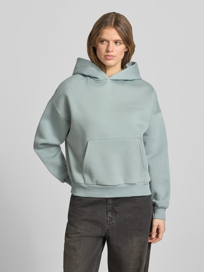 Frauenmodel trägt Hoodie mit Logo und Känguru-Tasche von Review in Hellblau, Baumwoll-Mix, Relaxed-Fit, Ganzkörperansicht von vorne.