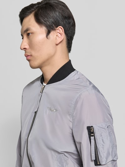 Männermodel trägt Bomberjacke mit Label-Patch und Eingrifftaschen von Original Bombers, hellgrau, Reißverschluss, Reißverschlusstasche, Nahaufnahme Oberkörper, Kontext: Herren, Gerader-Schnitt, Reißverschluss, Reißverschlusstasche am Ärmel, Eingrifftaschen mit Druckknopfverschluss, Label-Patch, Gerippte-Abschlüsse, Rückenlänge bei Größe M 67 cm, Schulterbreite bei Größe M 43 cm, Brustweite bei Größe M 53 cm, Ärmellänge bei Größe M 68 cm.
