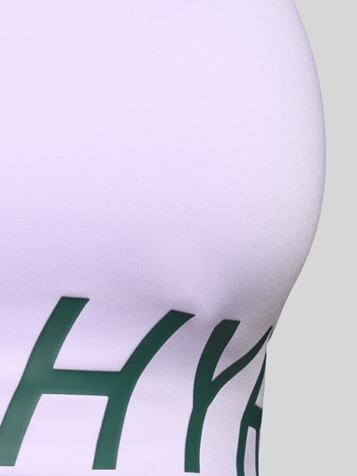 Vrouwenmodel draagt Sport-bh met labelprint van PUMA Performance, lichtpaars met groen detail, detailopname rechterborst.
