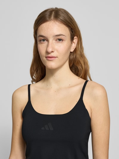 Frauenmodel trägt Tanktop mit Label-Print von adidas Sportswear in Schwarz, schlanke Passform, dehnbarer Material-Mix, Nahaufnahme Oberkörper.